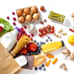 Top view of a paper bag full of canned food, fruits, vegetables, eggs, a milk bottle, berries, mushrooms, nuts, pasta, a chocolate bar and bread. The paper bag is laying on the white background and the food is coming out from it.